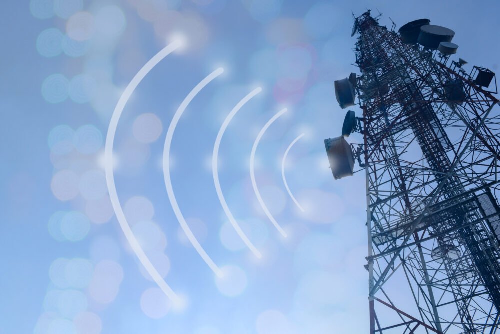 New Research Highlights Growing Health Risks of Cell Towers and EMF Exposure 1 Cell tower with wireless antennas against a blue sky and illustrated radiofrequency signal waves.